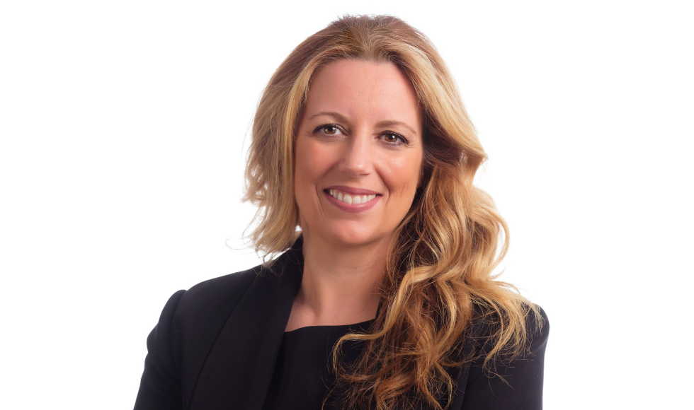 A person with blonde hair and a black blazer smiles to camera. The background is white.