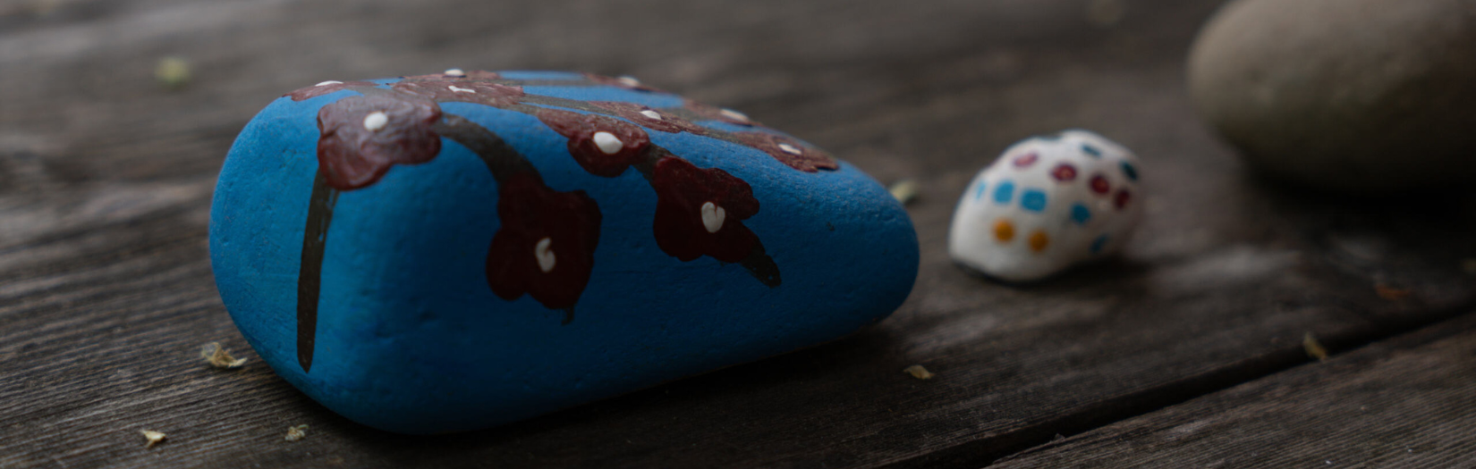 Three rocks sit on a wooden surface. One rock is blue and painted with a design, one is white and has small dots, and the third is grey.