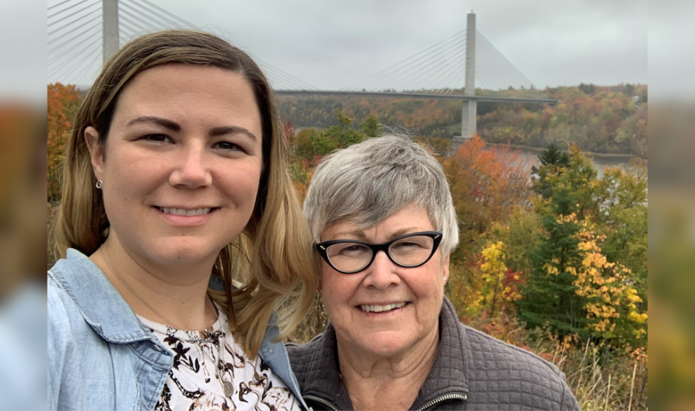 Two woman in outdoors in the fall, against a backdrop of fall colours and a bridge in the distance.