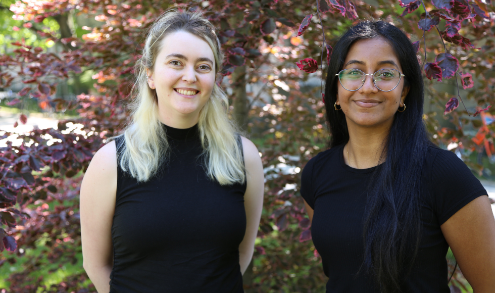 Two women stand beside each other, with a tree behind them.
