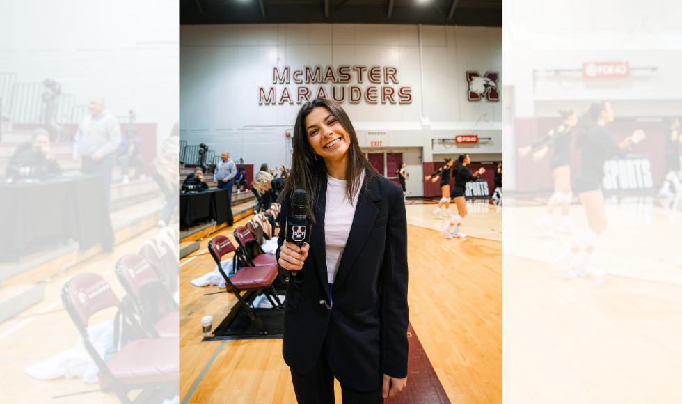 A woman holding a microphone stands on the side of a court, with a volleyball game going on behind her and the words 'McMaster Marauders' on the wall.