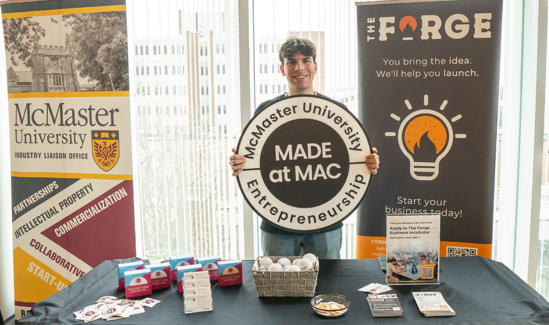 A student standing in front of a banner for the Forge and the McMaster Industry Liaison Office holds up a Made at Mac sign at a table during an entrepreneurship event.