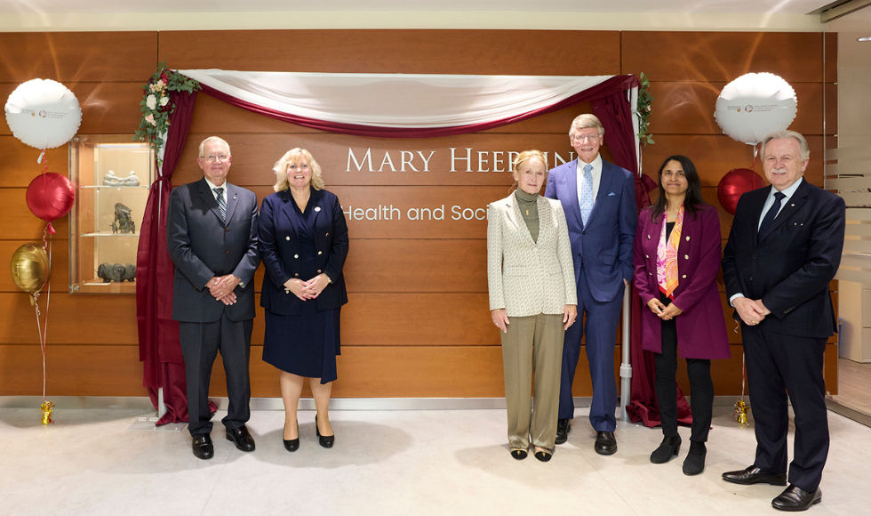 Six people in suits an dress clothes stand with balloons in front of a banner and wooden doors.