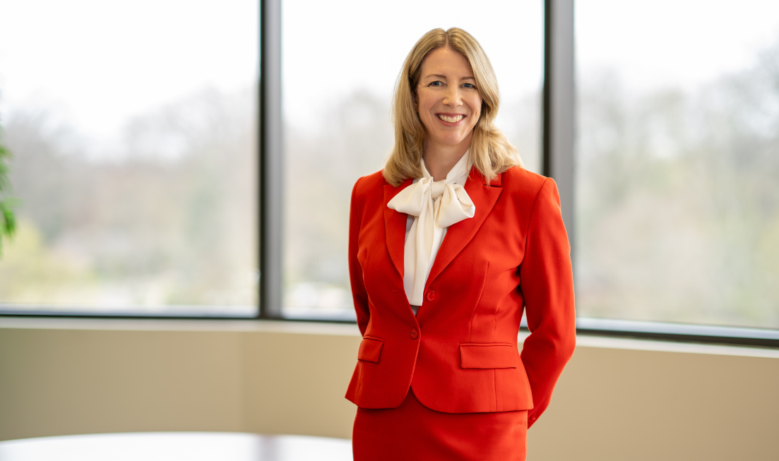 Maureen MacDonald, with shoulder-length blond hair, in a red suit with a white blouse, standing indoors in front of large windows.
