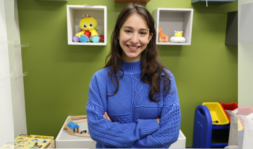 A woman in a blue sweater smiles and poses for the camera. Behind her, baby toys are visible on the ground and on shelving units.
