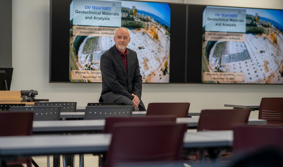 A man in a suit sits at the front of an empty classroom.