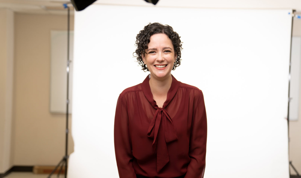 A woman in a red shirt stands in front of a white backdrop in a photo studio.