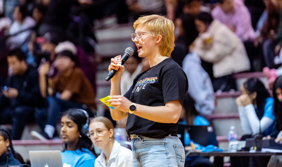 A woman stands at the front of several rows of bleachers, speaking into a microphone, holding a small note in her other hand.
