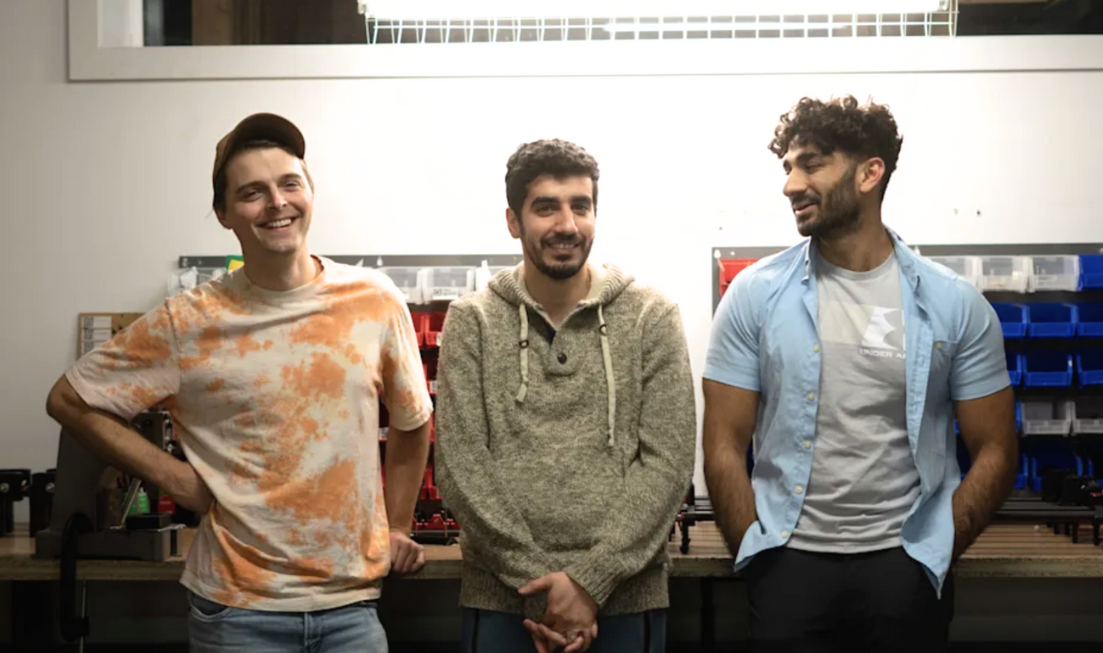 Three smiling young men standing beside each other leaning against a work surface.