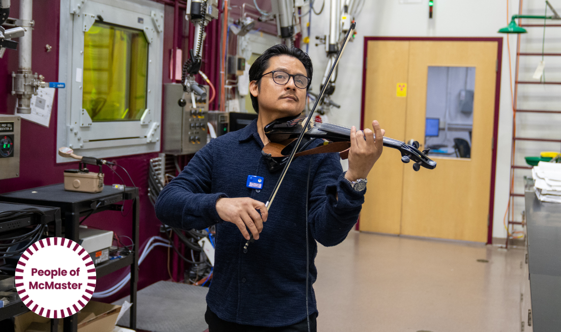 Alain Pamuce, a dark-haired man with glasses in dark blue clothes, plays the violin in the hot cells of the Tandem Accelerator building at McMaster.