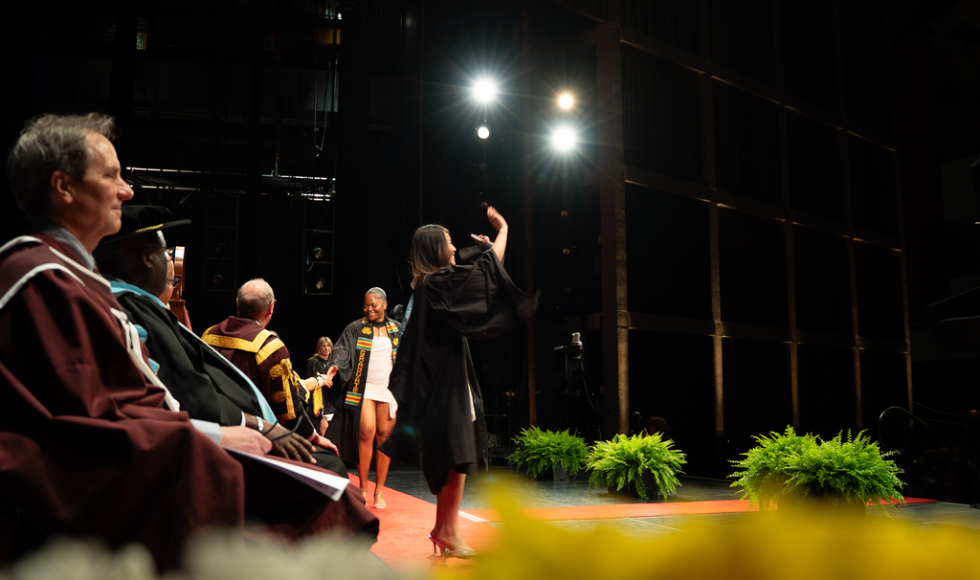At a graduation ceremony, a woman stops on the stage to wave to the crowd.