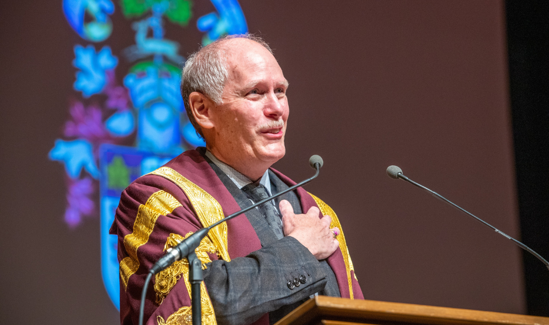 David Farrar, in McMaster University President's ceremonial robe, places his hand on his heart and smiles as he speaks to graduating students on stage at convocation.