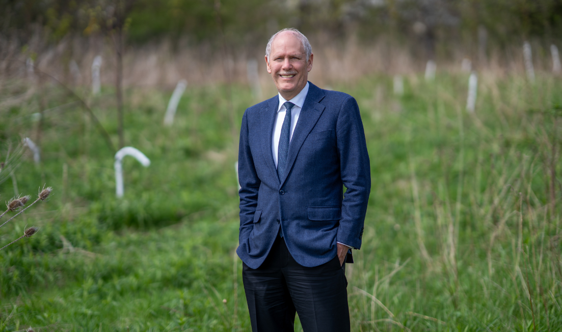 President David Farrar wearing a blue suit and tie with a white shirt, standing with his hands in his pockets against a backdrop of greenery, smiling.