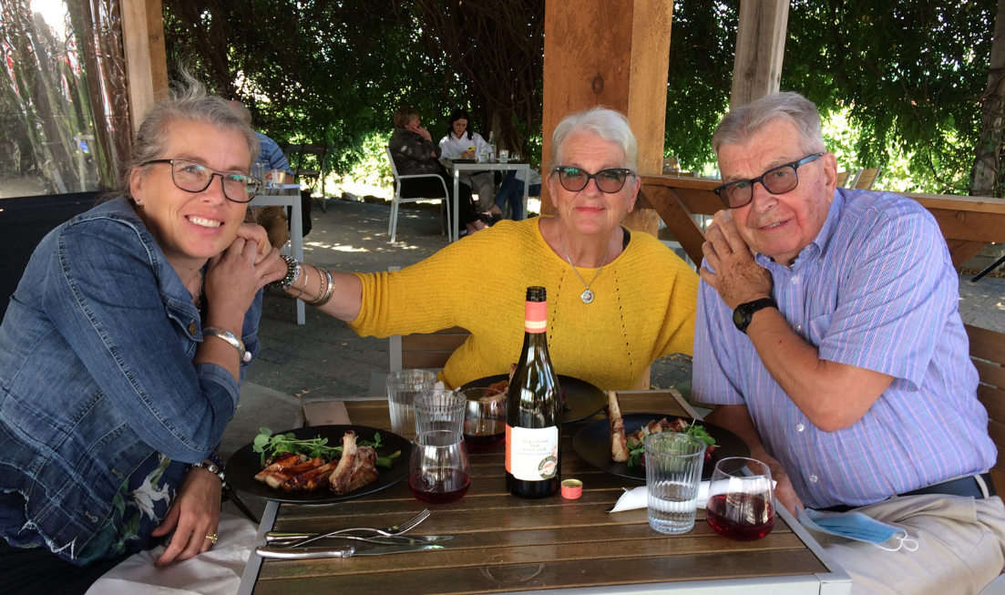 Three smiling people holding each others hands as they at a table outdoors with the remains of a meal on it.