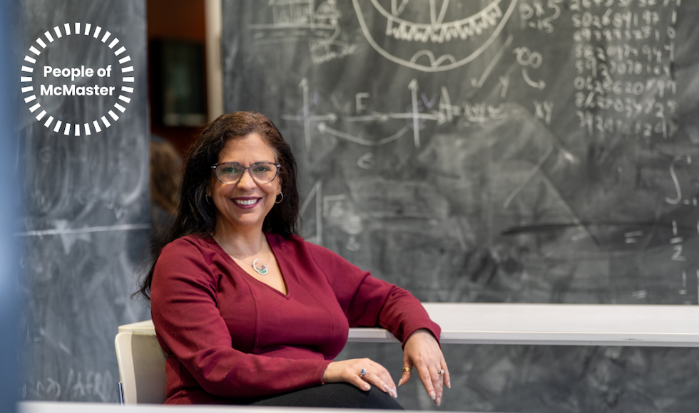 A women in a red sweater sits at a table in front of a chalkboard, smiling towards the camera.
