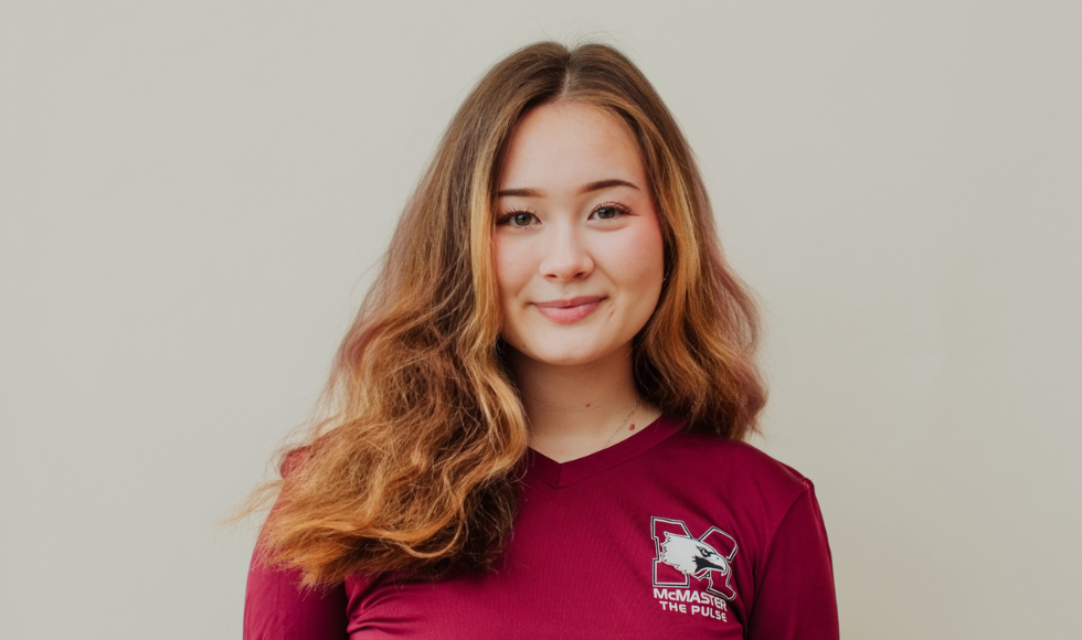 A headshot of a young woman, who is wearing a red long-sleeved shirt. The shirt has a small logo on the front that reads 'McMaster, the Pulse,' with a graphic of a bird and the letter M.