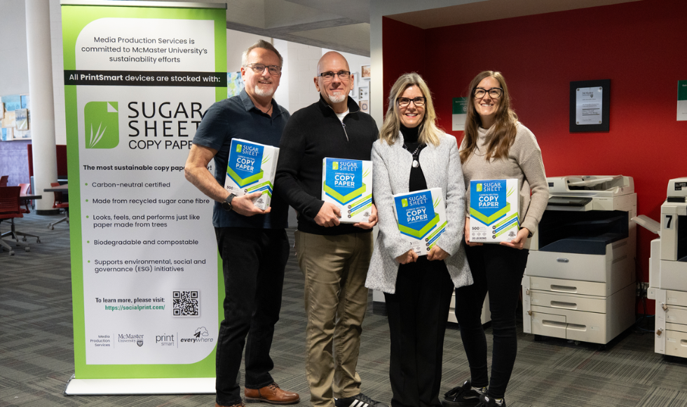 Four people, each holding a ream of printer paper, stand beside a printer.