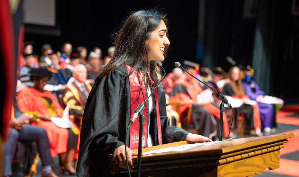 A young woman in graduation robes stands at a podium on a stage, looking out towards the theatre and speaking. Several people wearing robes are sitting on the stage behind her.