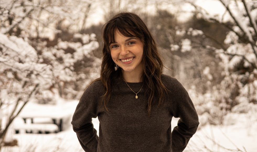 A headshot of a woman in a dark sweater, standing in front of a snowybackground.