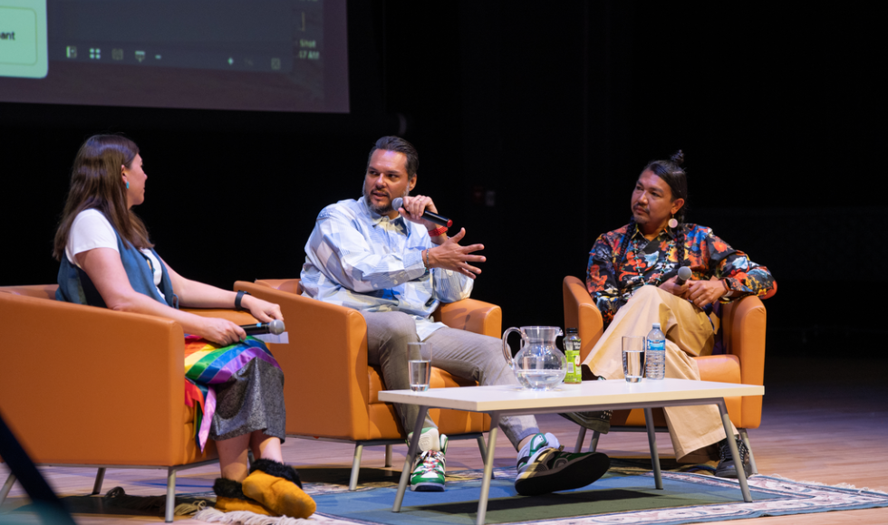 On a stage, three people sit in armchairs, with a coffee table in front of them. They are seated in a semi-circle, with the person in the middle speaking into a microphone.