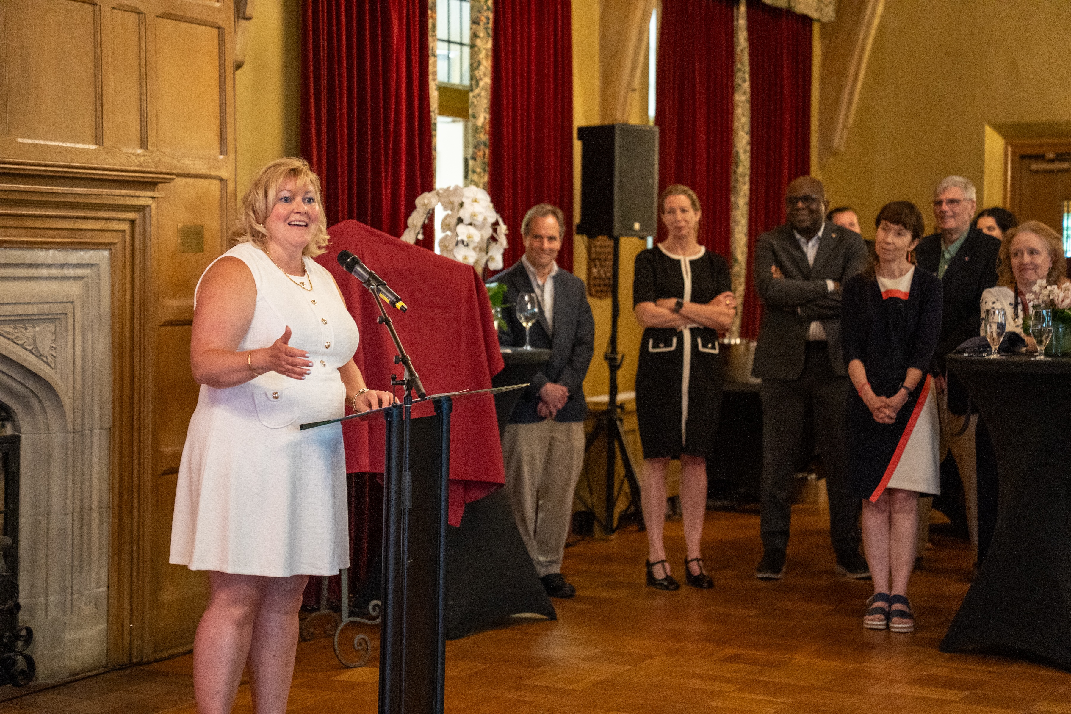 Susan Tighe speaks into a mic, watched by jerry Hurley, Maureen MacDonald, Bonny Ibhawoh and Vivian Lewis, during a gathering in Alumni Memorial Hall