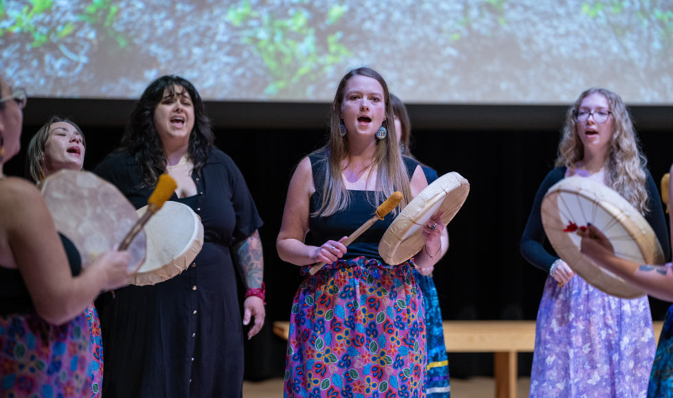Several women with drums stand in a circle, singing.