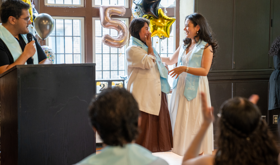 Two women in dress clothes stand on stage, looking at one another. They are wearing blue graduation stoles.
