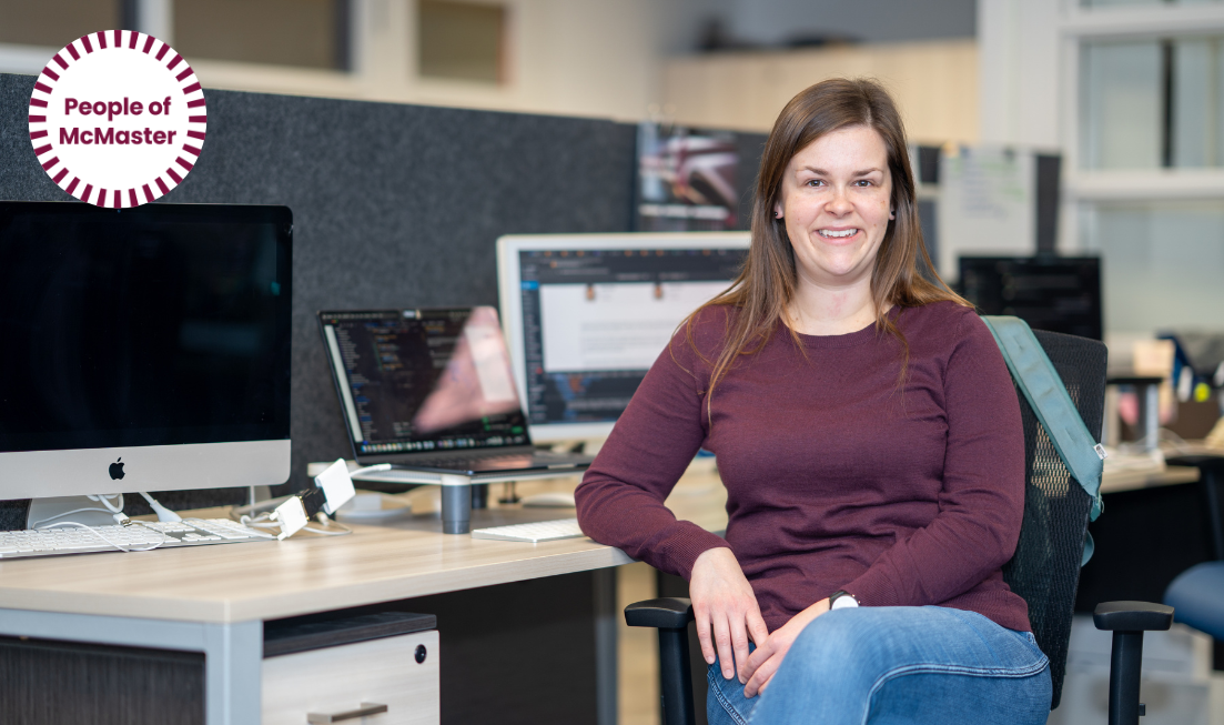 Smiling Melanie Phillips — a woman with long brown hair, a maroon top and blue jeans — sitting at a desk with a laptop and a few screens.