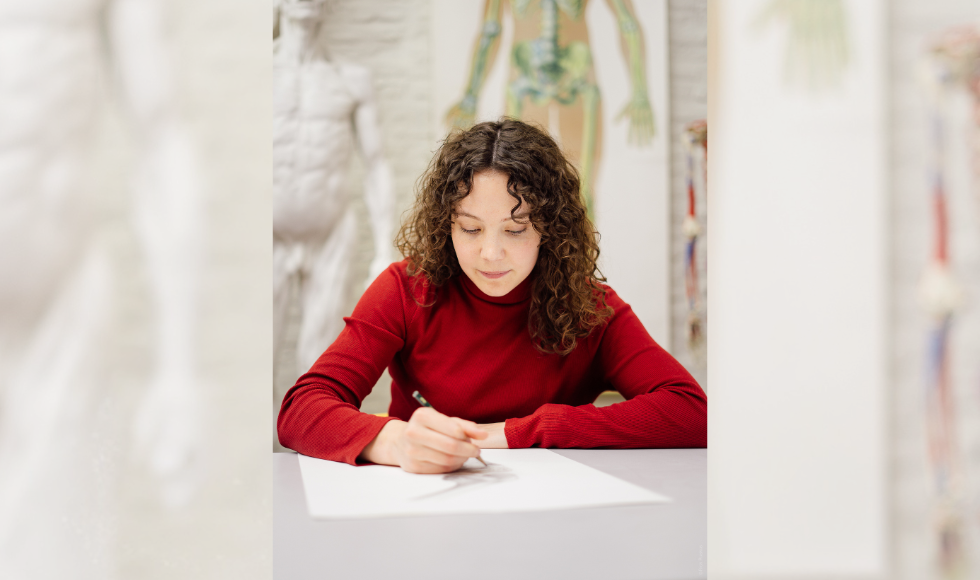 A woman sits at a table, drawing, with anatomical posters on the wall behind her.