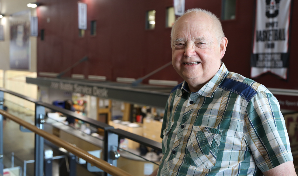 An older man stands in front of a railing, with a gym entryway behind him.
