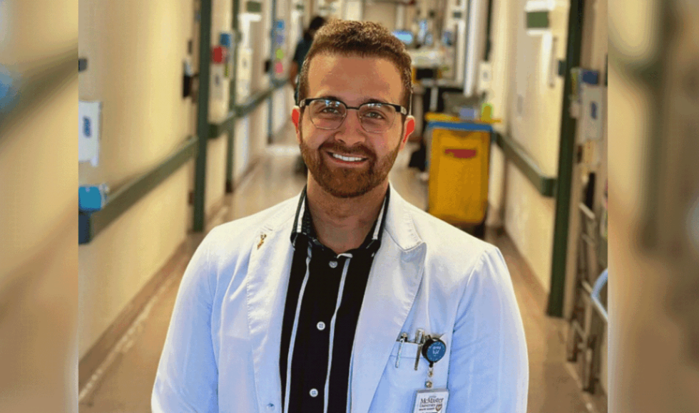 A man in a white lab coat stands in a hospital hallway, smiling.