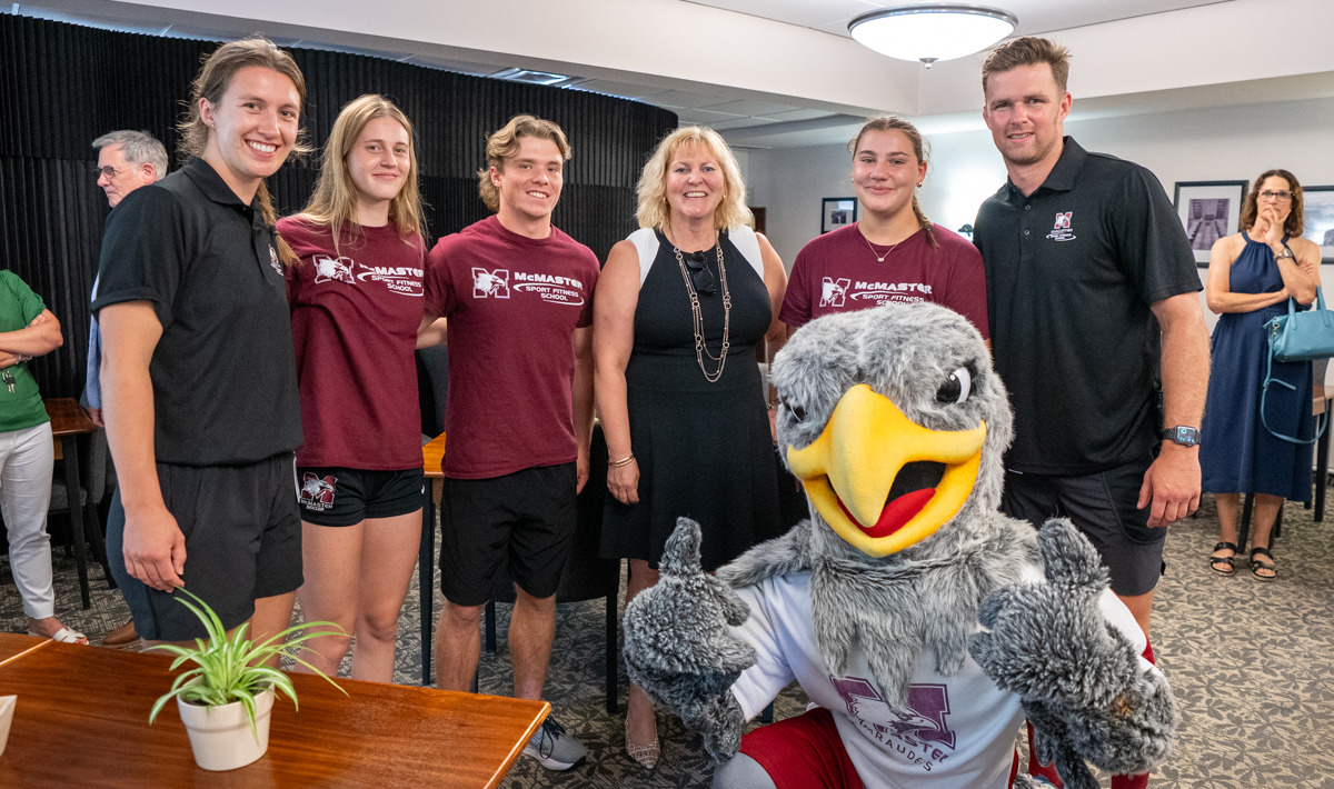 Mascot Mac the Marauder kneels and gives a thumbs up, with five student athletes and Susan Tighe standing behind him, all smiling, at a gathering indoors.