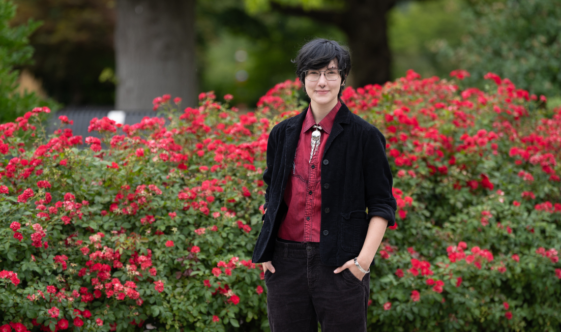 A dark-haired man with glasses, wearing maroon and black, standing in front of a hedge of red rose bushes in bloom.