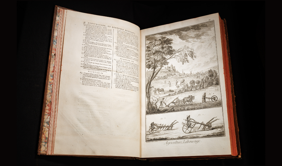 A large, old book lies open on a black background. The book has text on one open page, and a large illustration of an agricultural scene on the other open page.