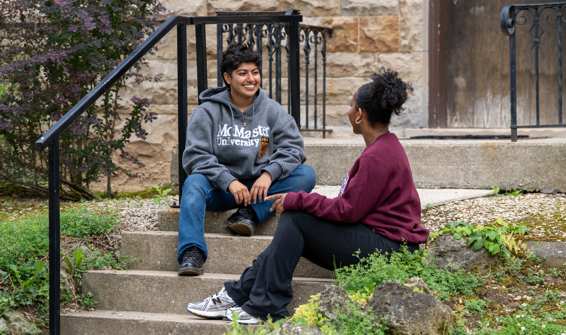 A smiling student in a McMaster hoodie sits on a step, chatting with a second student in a hoodie, whose back is to the camera.
