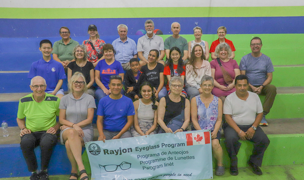 A group shot of about thirty people, sitting in three rows, smiling. The people in the front row are holding up a large banner with a Canadian flag, a picture of eyeglasses, and text on it.