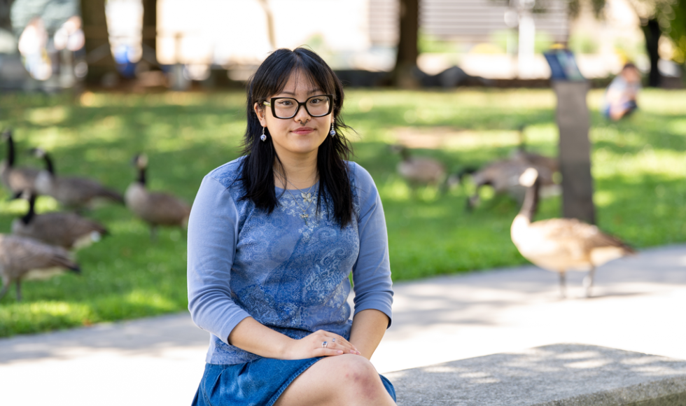A woman sits on a bench outdoors, with several geese in the background behind her.