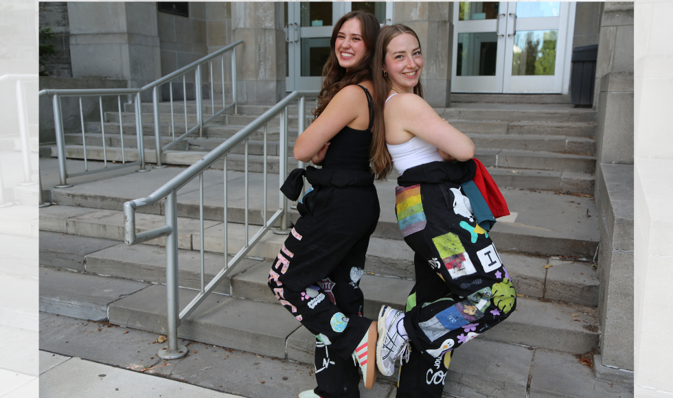 Two women pose back-to-back on a set of concrete steps. They are each wearing a black jumpsuit covered in colourful patches.