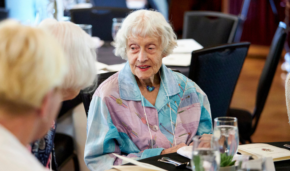 An older woman sits at a table, talking to people.