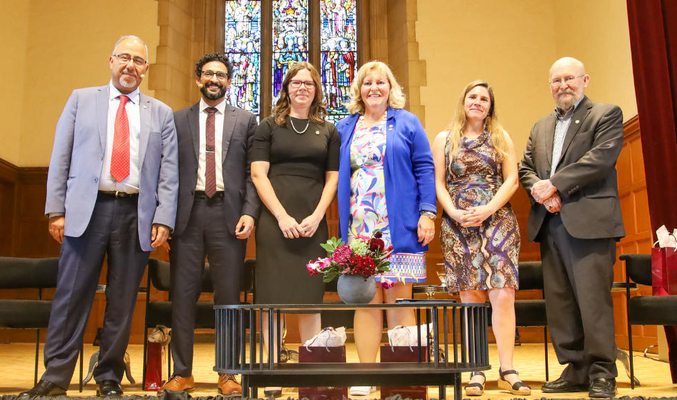 Six people standing in a row on the stage in Convocation Hall smile down at the camera.
