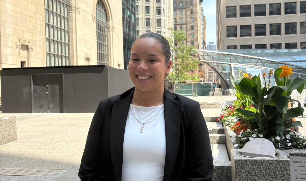 A woman in a blazer stands outside large stone buildings in the city.