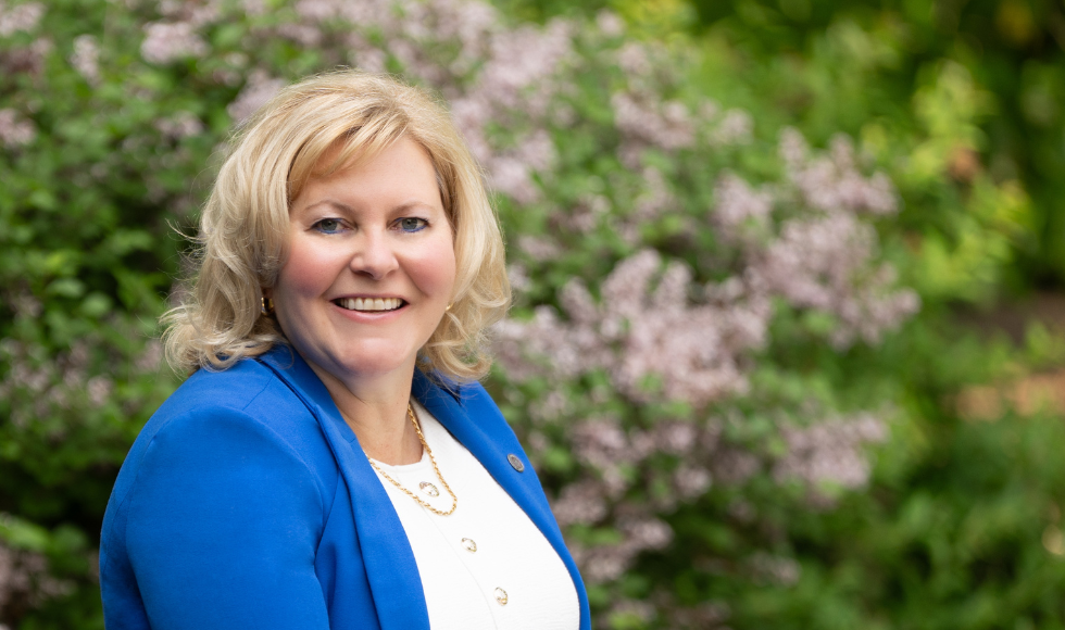 Susan Tighe, president and vice-chancellor of McMaster University, faces camera in a blue jacket. Purple flowers behind her.