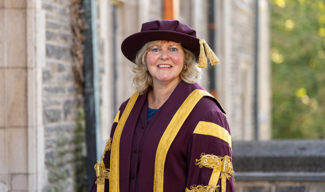 Susan Tighe, smiling, wearing the McMaster presidential robes and hat against a backdrop of a campus building.