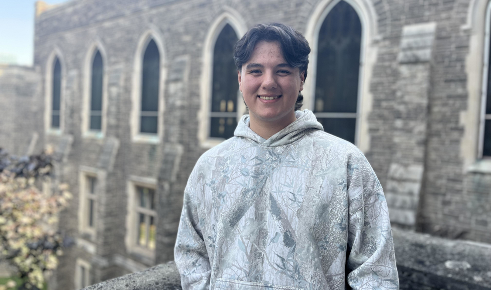 A young man in a hoodie stands in front of a grey stone building.