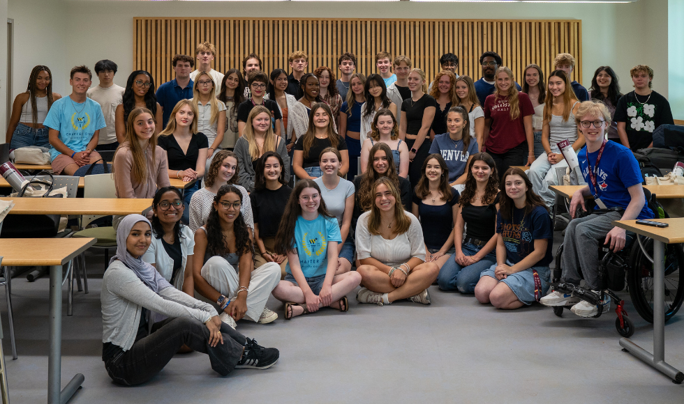 A group of about 50 smiling students standing and sitting in rows in a classroom with the desks moved out of the way for a group picture.